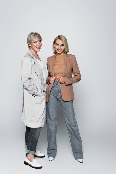 full length view of mother and daughter in stylish outfit smiling at camera on grey - Foto, afbeelding