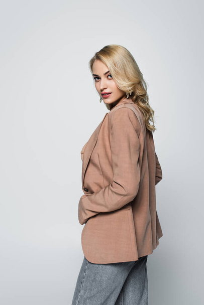 blonde, sensual woman in beige blazer looking at camera isolated on grey - 写真・画像
