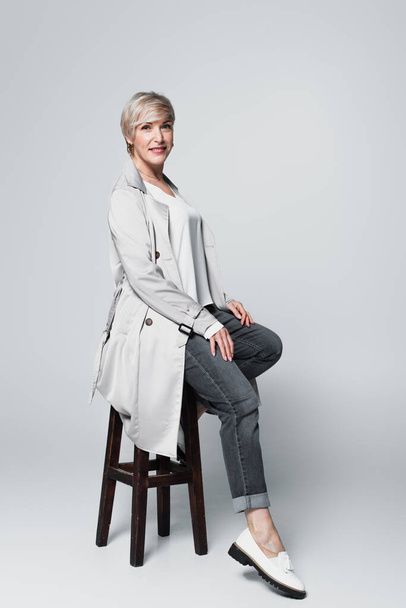 ash-blonde woman in trendy autumn outfit sitting on high stool on grey - Foto, Imagen