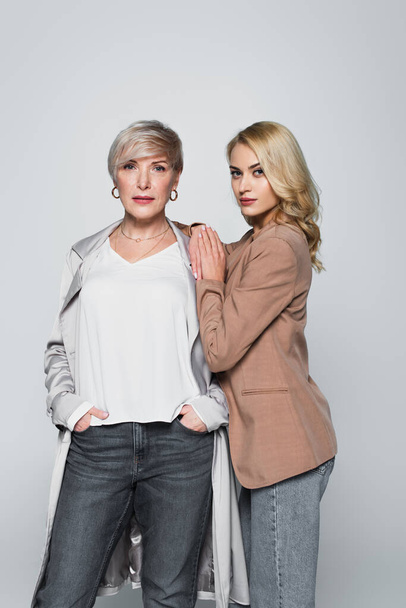 young woman touching shoulder of stylish mother while looking at camera isolated on grey - Photo, image
