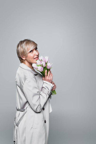 Frau mittleren Alters im grauen Trenchcoat posiert mit frischen Tulpen isoliert auf grau - Foto, Bild