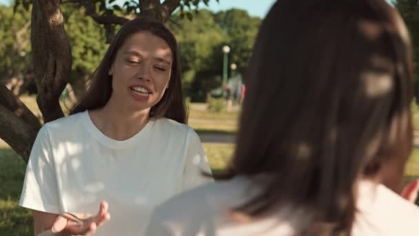 Olkapään yli vyötärö-up onnellinen nuori valkoihoinen nainen valkoinen t-paita istuu puussa puistossa, hymyillen, chattailuun tunnistamaton ystävä - Materiaali, video
