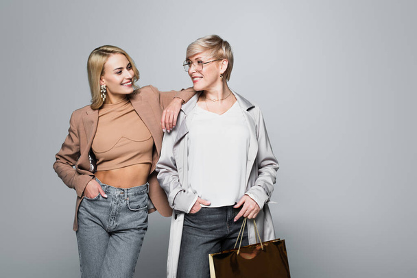 trendy mother and daughter smiling at each other while posing with hands in pockets on grey - Foto, afbeelding