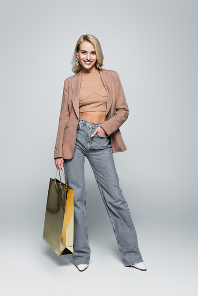 full length view of fashionable woman posing with shiny shopping bag on grey - Foto, Imagem