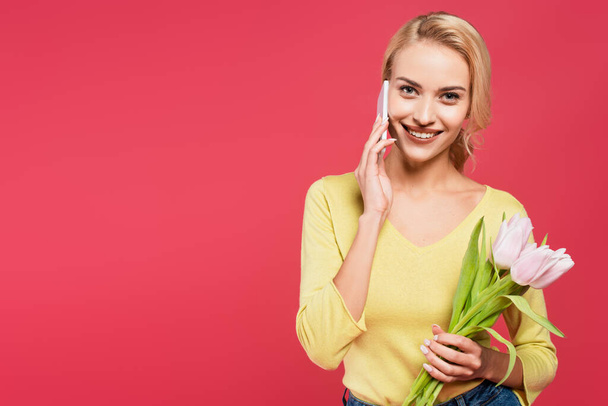 blonde woman in yellow jumper talking on cellphone while holding tulips isolated on pink - Foto, Imagem