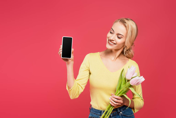 fröhliche, blonde Frau posiert mit Handy und Tulpen isoliert auf rosa - Foto, Bild