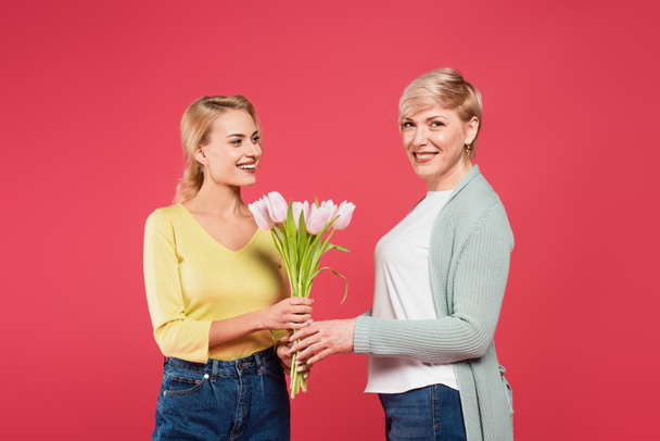 smiling woman presenting tulips to happy mother looking at camera isolated on pink - Photo, Image