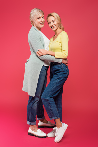 full length view of cheerful mother and daughter in stylish clothes embracing on pink - Foto, afbeelding