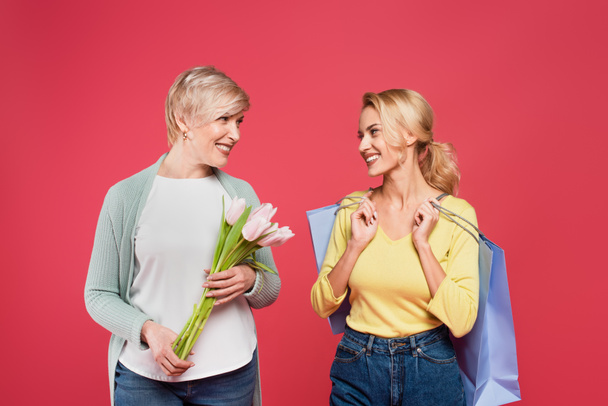 cheerful mother and daughter with tulips and shopping bags smiling at each other isolated on pink - Photo, image