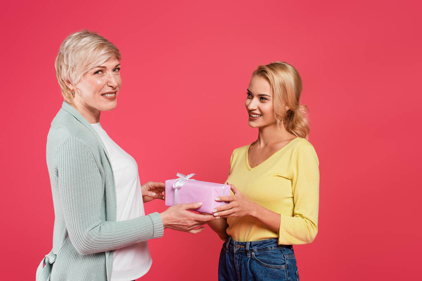 happy middle aged woman taking gift from adult daughter while looking at camera isolated on pink - 写真・画像