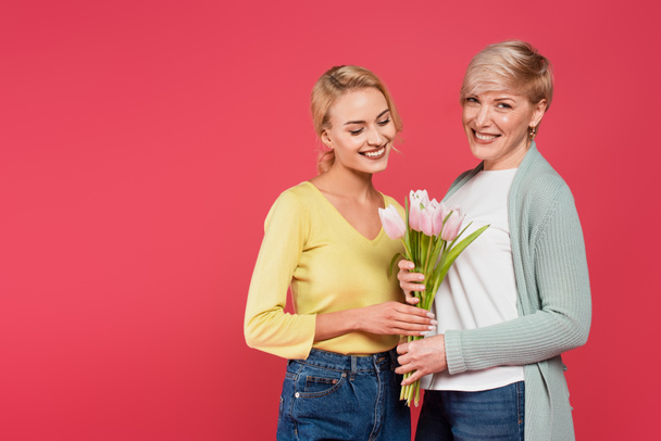 веселые, стильные мать и дочь держа тюльпаны изолированы на розовый - Фото, изображение