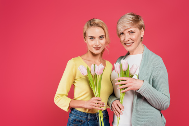 stylish, joyful mother and daughter holding tulips and looking at camera isolated on pink - Fotó, kép