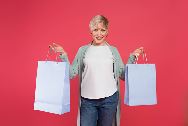 Счастливая блондинка, держа в руках сумки с покупками, глядя в камеру, изолированную от розового - Фото, изображение