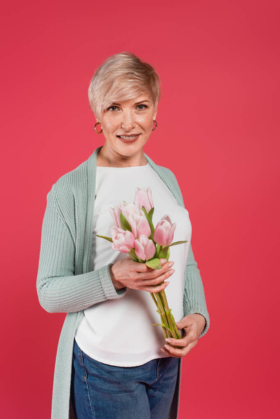 зрелая, стильная женщина, держащая свежие тюльпаны, глядя на камеру, изолированную на розовый - Фото, изображение