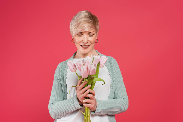 веселая женщина средних лет, держащая свежие тюльпаны изолированы на розовый - Фото, изображение