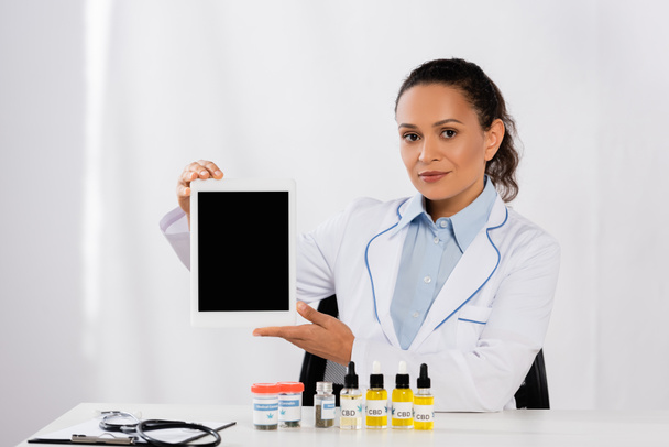 Afrikaans-Amerikaanse arts in witte jas met behulp van digitale tablet met blanco scherm in de buurt van flessen met medische cannabis  - Foto, afbeelding