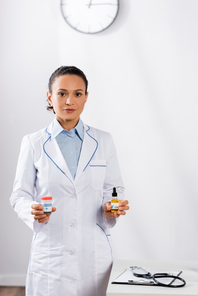 Afrikaans-Amerikaanse arts in witte jas met flessen met cbd en medicinale cannabis  - Foto, afbeelding
