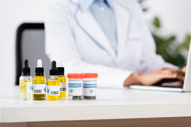 bottles with medical cannabis and cbd lettering on table near doctor on blurred background - Photo, Image