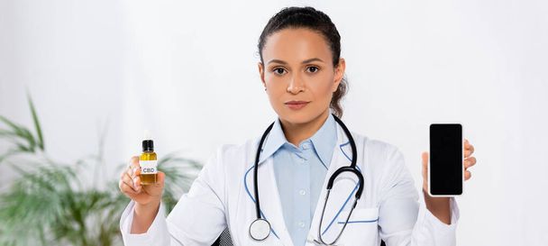 Африканский американский врач держит бутылку с CBD и смартфон с пустым экраном, баннер - Фото, изображение