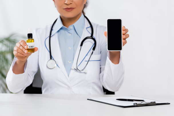 обрезанный вид африканского американского врача, держащего бутылку с CBD и смартфон с пустым экраном - Фото, изображение