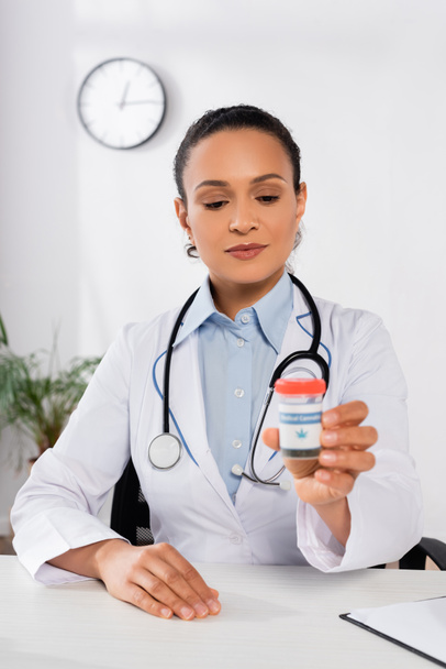 Африканский американский врач со стетоскопом, держащий бутылку с медицинскими надписями на конопле в клинике - Фото, изображение