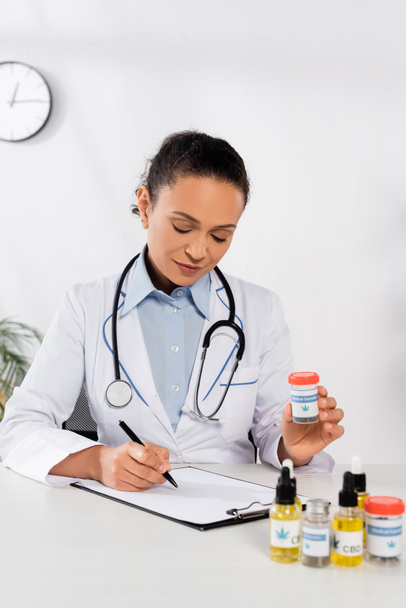 bruna medico afroamericano con stetoscopio bottiglia con scritte di cannabis medica e scrittura sugli appunti - Foto, immagini