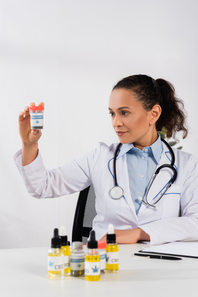 brunette african american woman with stethoscope looking at bottle with medical cannabis lettering in clinic - Photo, Image