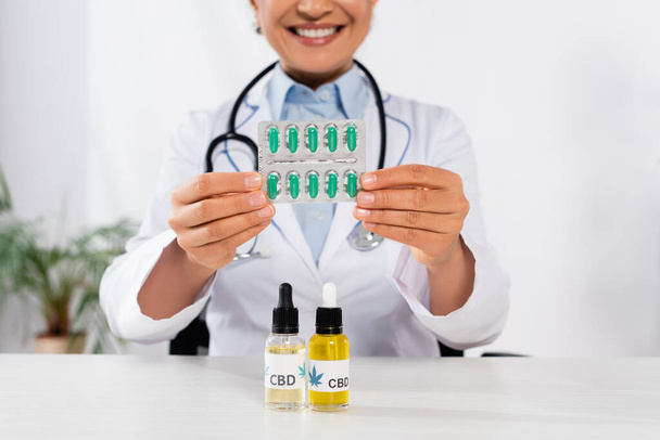 bijgesneden weergave van vrolijke Afrikaanse Amerikaanse arts met stethoscoop met blisterverpakking met capsules in de buurt van cbd flessen op bureau - Foto, afbeelding