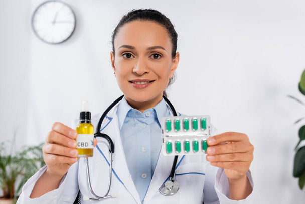 médecin afro-américain joyeux avec stéthoscope tenant blister paquet avec capsules près de bouteille cbd - Photo, image
