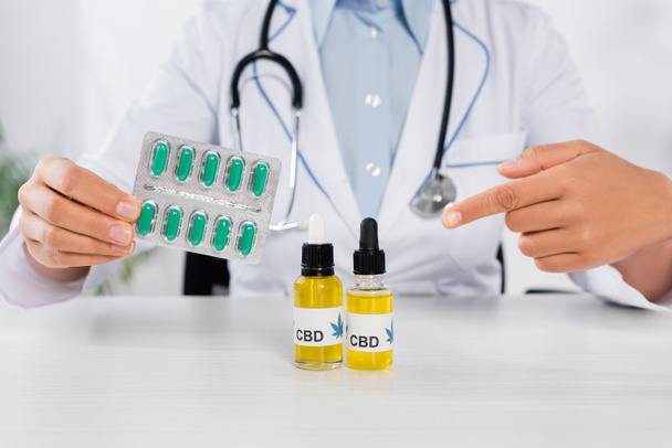 cropped view of doctor with stethoscope pointing with finger at cbd bottles while holding blister pack with capsules  - Photo, Image