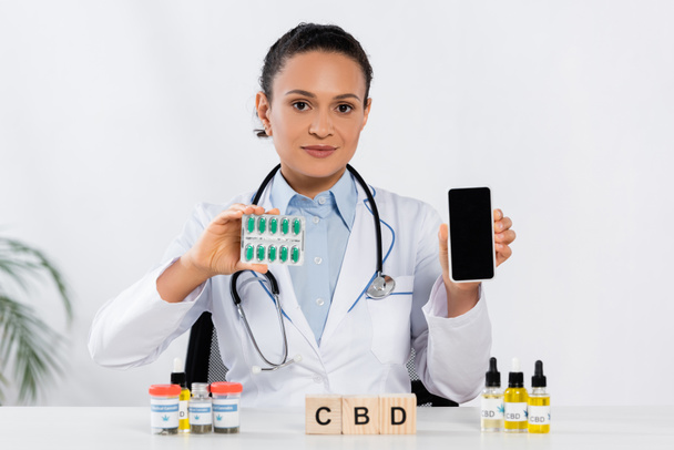 médecin afro-américain avec stéthoscope tenant plaquette thermoformée avec capsules et smartphone près de cubes et bouteilles cbd  - Photo, image