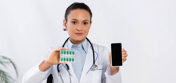 Afrikaans-Amerikaanse arts met stethoscoop met blisterverpakking met capsules en smartphone met blanco scherm, banner - Foto, afbeelding
