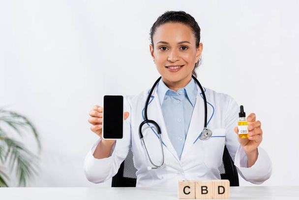 doctor afroamericano feliz sosteniendo la botella y el teléfono inteligente con la pantalla en blanco cerca de cubos del cbd  - Foto, Imagen