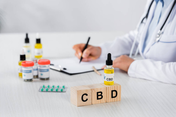 частичный взгляд врача, выписывающего рецепт рядом с кубиками CBD и лекарствами на столе - Фото, изображение