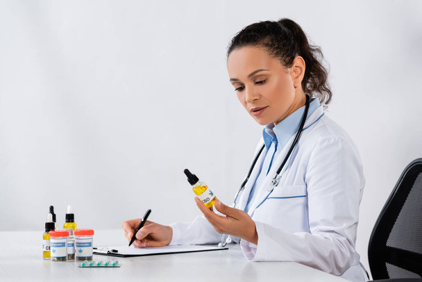 Africano médico americano que escreve prescrição e segurando garrafa cbd perto de medicação na mesa - Foto, Imagem