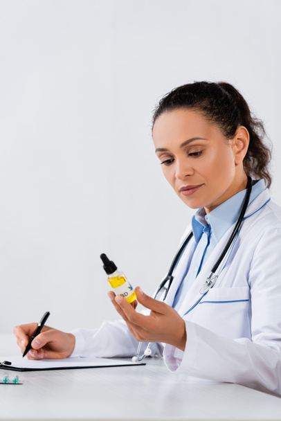 médico afroamericano escribiendo prescripción y sosteniendo botella de cbd  - Foto, Imagen