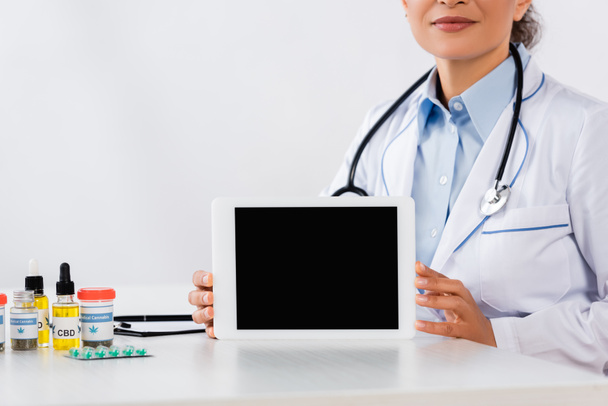 vista parziale del medico afro-americano con tablet digitale con schermo bianco vicino ai farmaci per la cannabis sulla scrivania - Foto, immagini