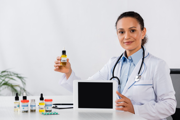 Mutlu Afrikalı Amerikalı doktor elinde boş ekranlı dijital tablet ve ilaç masasının yanında cbd şişesi tutuyor. - Fotoğraf, Görsel