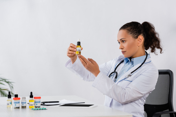 medico afroamericano in possesso di bottiglia cbd vicino a farmaci e tablet digitale sulla scrivania - Foto, immagini