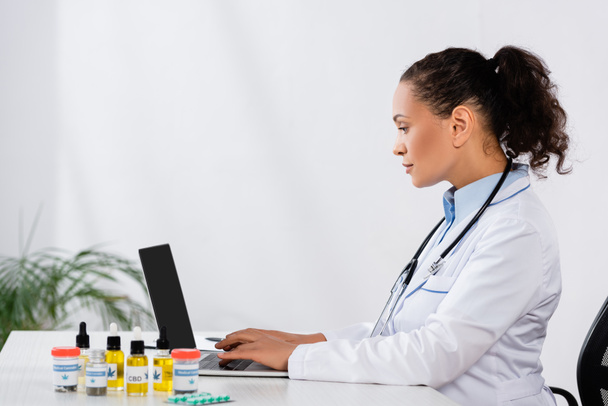 vista laterale del medico afro-americano che digita sul computer portatile vicino ai farmaci per la cannabis sulla scrivania - Foto, immagini