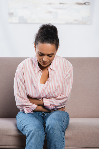 mujer afroamericana que sufre de dolor abdominal mientras está sentada en el sofá en la sala de estar  - Foto, imagen