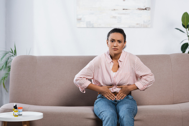 afro-amerikai nő szenved hasi fájdalom, miközben ül a kanapén a nappaliban  - Fotó, kép