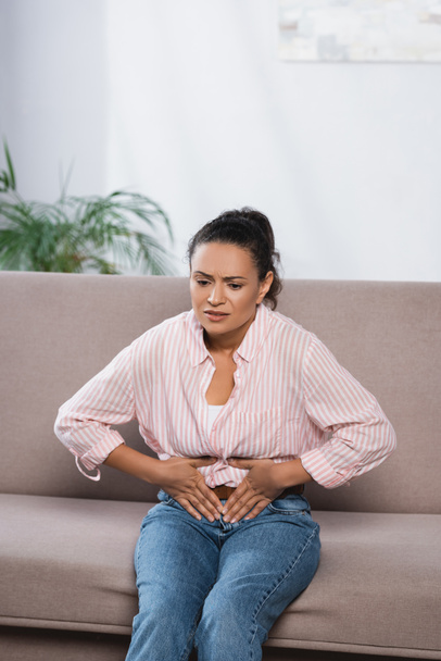 femme afro-américaine souffrant de douleurs abdominales tout en étant assis sur le canapé - Photo, image