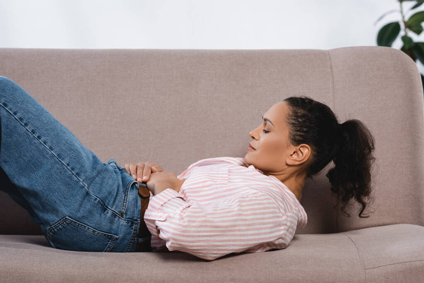 Afrikaans amerikaanse vrouw die lijden aan maagpijn terwijl liggend op de bank in de woonkamer  - Foto, afbeelding