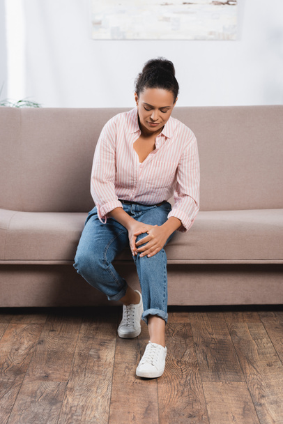 Afričanky americká žena trpí zezadu v koleni, zatímco sedí na gauči - Fotografie, Obrázek