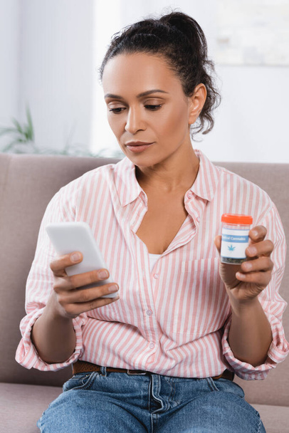 афроамериканська жінка тримає пляшку з медичним канабісом і дивиться на смартфон у вітальні
 - Фото, зображення