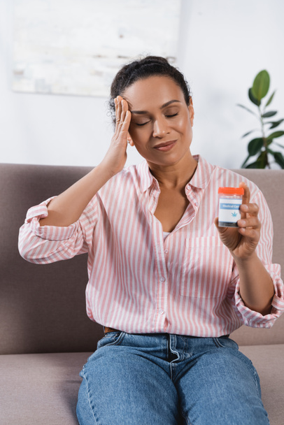 african american woman with closed eyes suffering from migraine and holding bottle with medical cannabis  - Photo, Image
