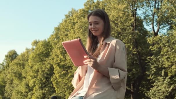 Средний снимок молодой радостной белой женщины, улыбающейся, сидящей на переднем плане высоких зеленых деревьев на открытом воздухе, с помощью планшетного компьютера в розовом чехле в солнечный день - Кадры, видео