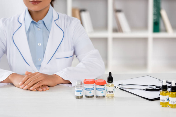 обрізаний вид на афро-американського лікаря в білому пальто біля пляшок з медичним канабісом і КДБ на столі
 - Фото, зображення