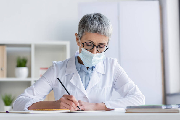 Tıbbi maskeli olgun bir doktor bulanık arkaplanda deftere yazıyor.  - Fotoğraf, Görsel